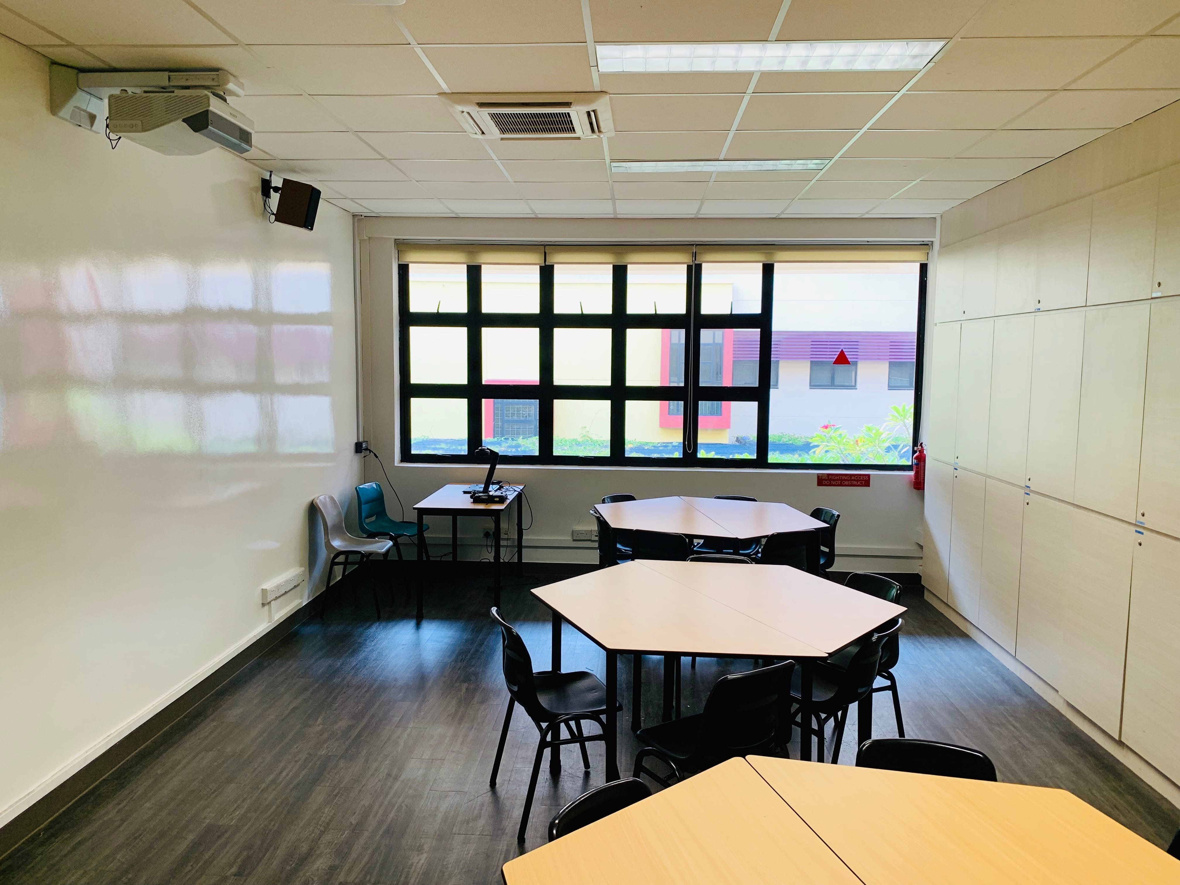 Empty classroom with hexagonal group tables, black chairs, a whiteboard, projector, and large windows overlooking a building.